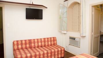 Ein Zimmer mit einer Couch, einem an der Wand montierten Fernseher, einem Fenster mit Rollläden und einer Tür zu einem anderen Raum.