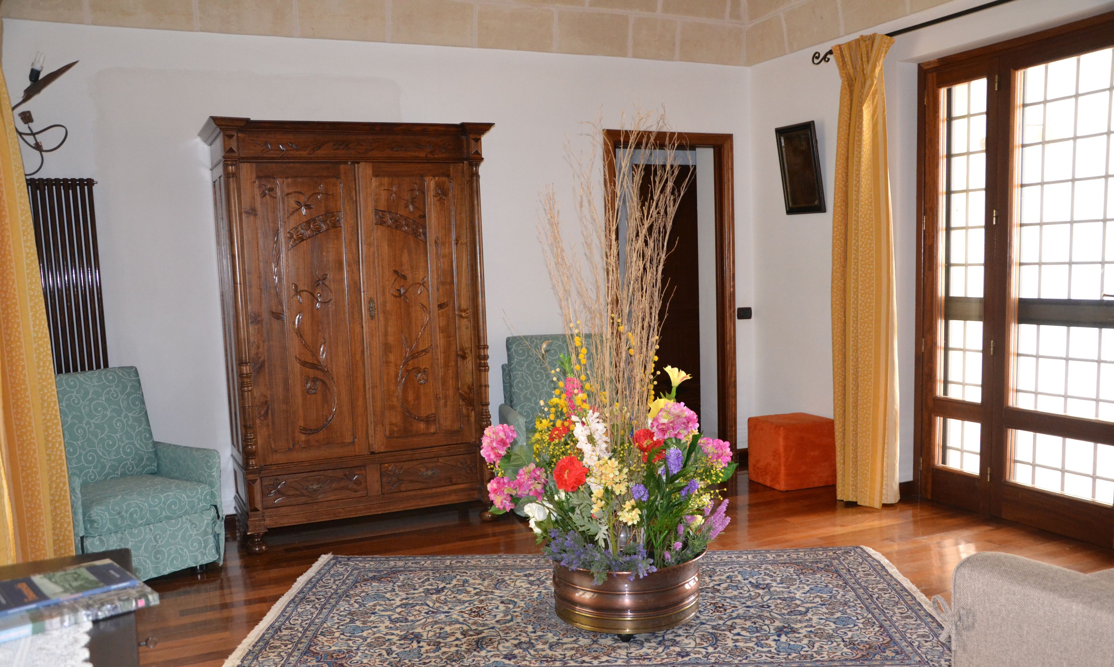 A room with a wooden wardrobe, a green armchair, a metal vase with flowers, and a patterned rug.