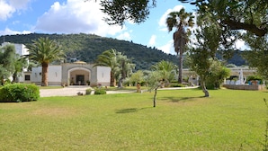 Um edifício com fachada de pedra, entrada em arco e um jardim bem cuidado com palmeiras e arbustos.