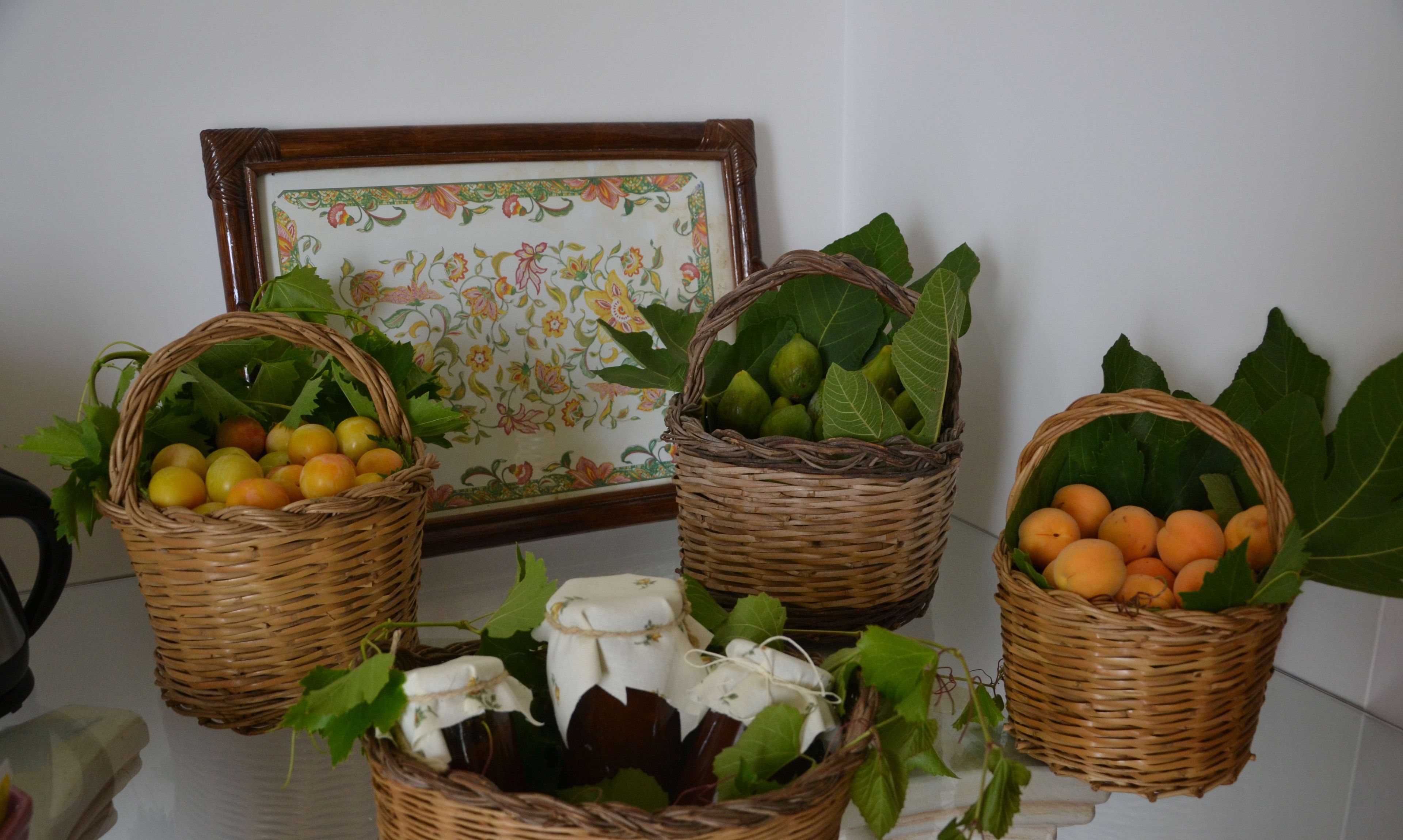 Trois paniers en osier remplis de fruits et de légumes, un tableau encadré orné d’une bordure florale et un mur recouvert de carreaux blancs.
