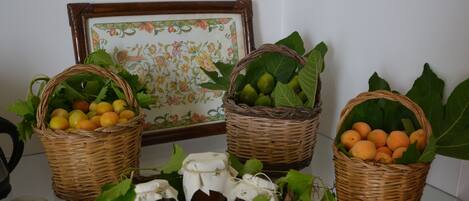 Trois paniers en osier remplis de fruits et de légumes, un tableau encadré orné d’une bordure florale et un mur recouvert de carreaux blancs.