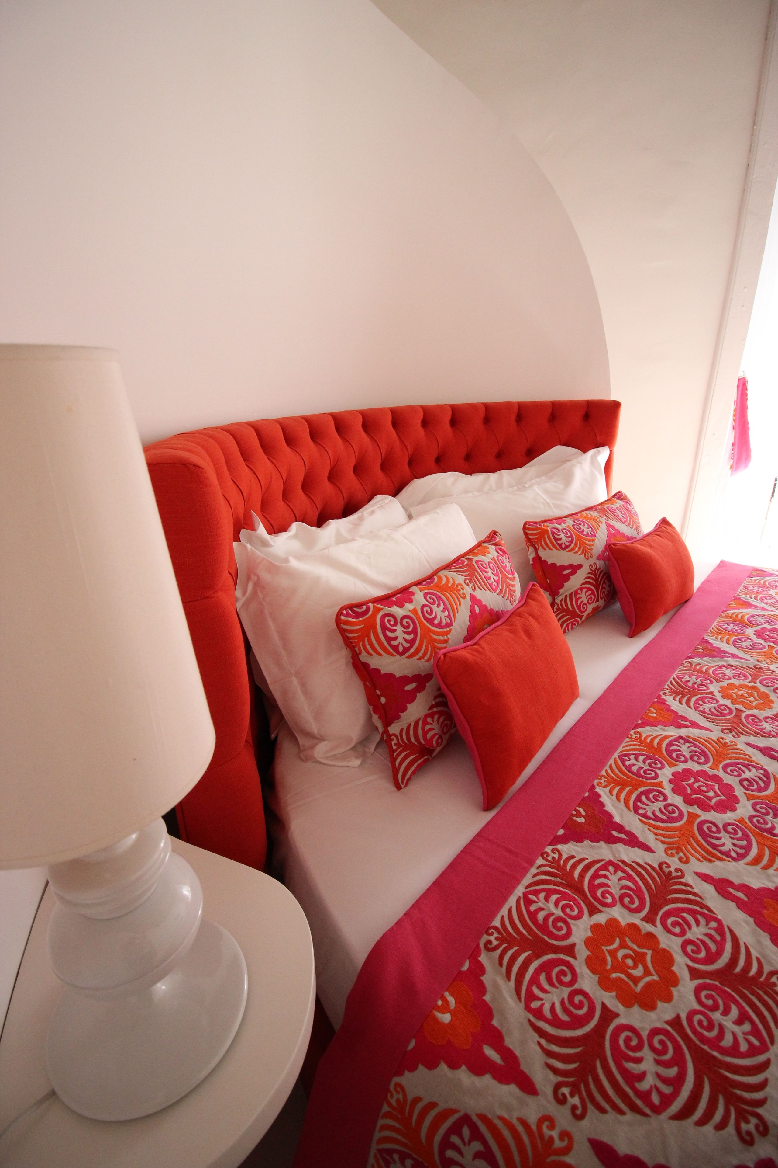 Une chambre à coucher avec une tête de lit capitonnée, du linge de lit blanc et une couvre-lit aux motifs rouges et oranges.
