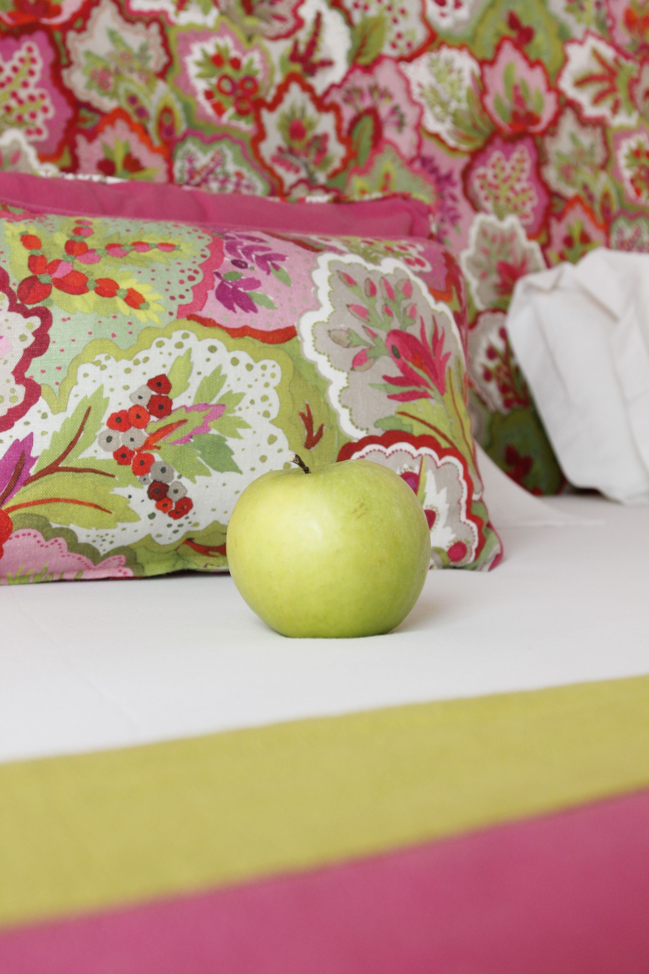 Une pomme verte posée sur un lit agrémenté d’un oreiller à motifs floraux et d’une couverture blanche.