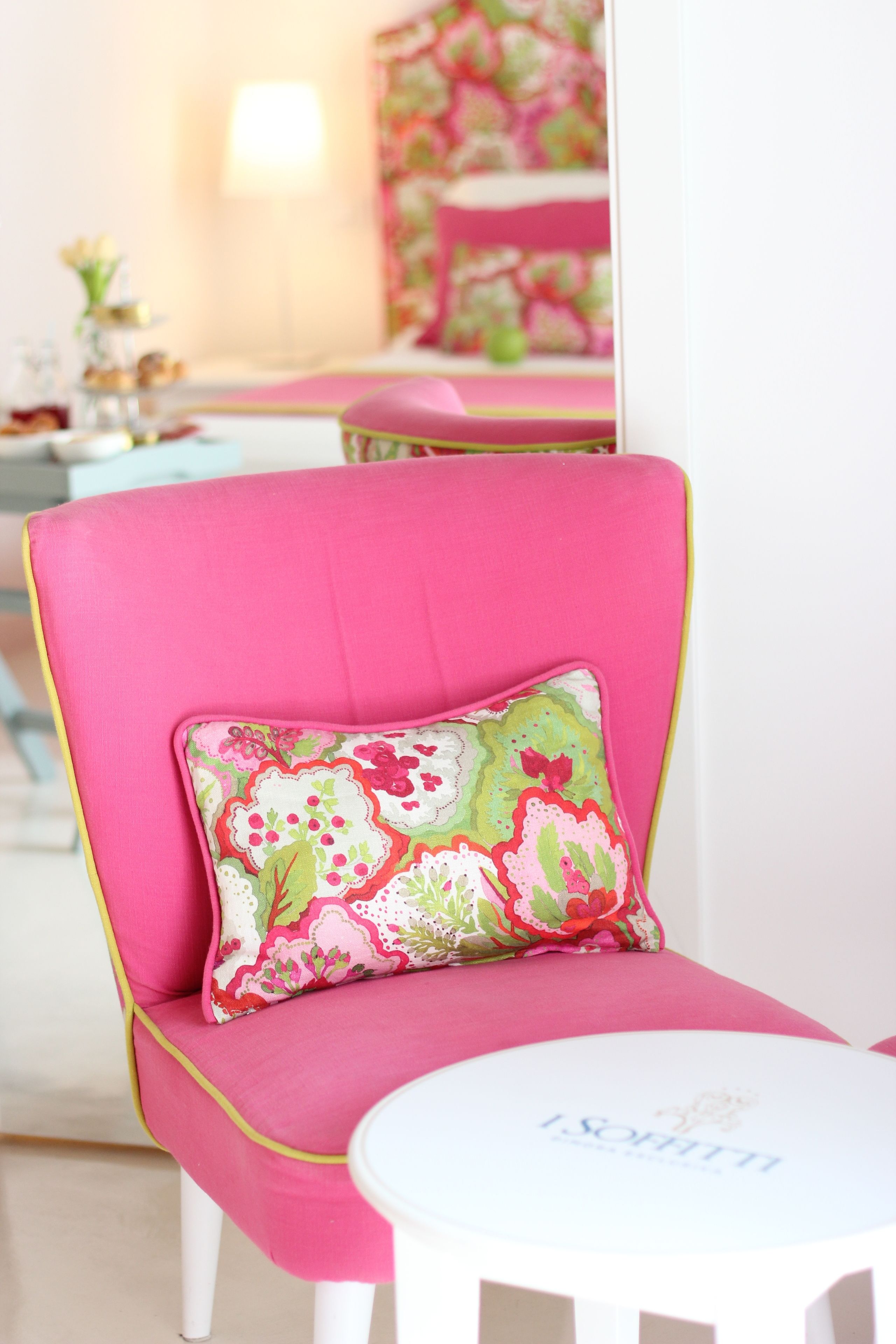 A pink chair with a floral pillow, a white table with a nameplate, and a mirror reflecting a room with pink and floral decor.