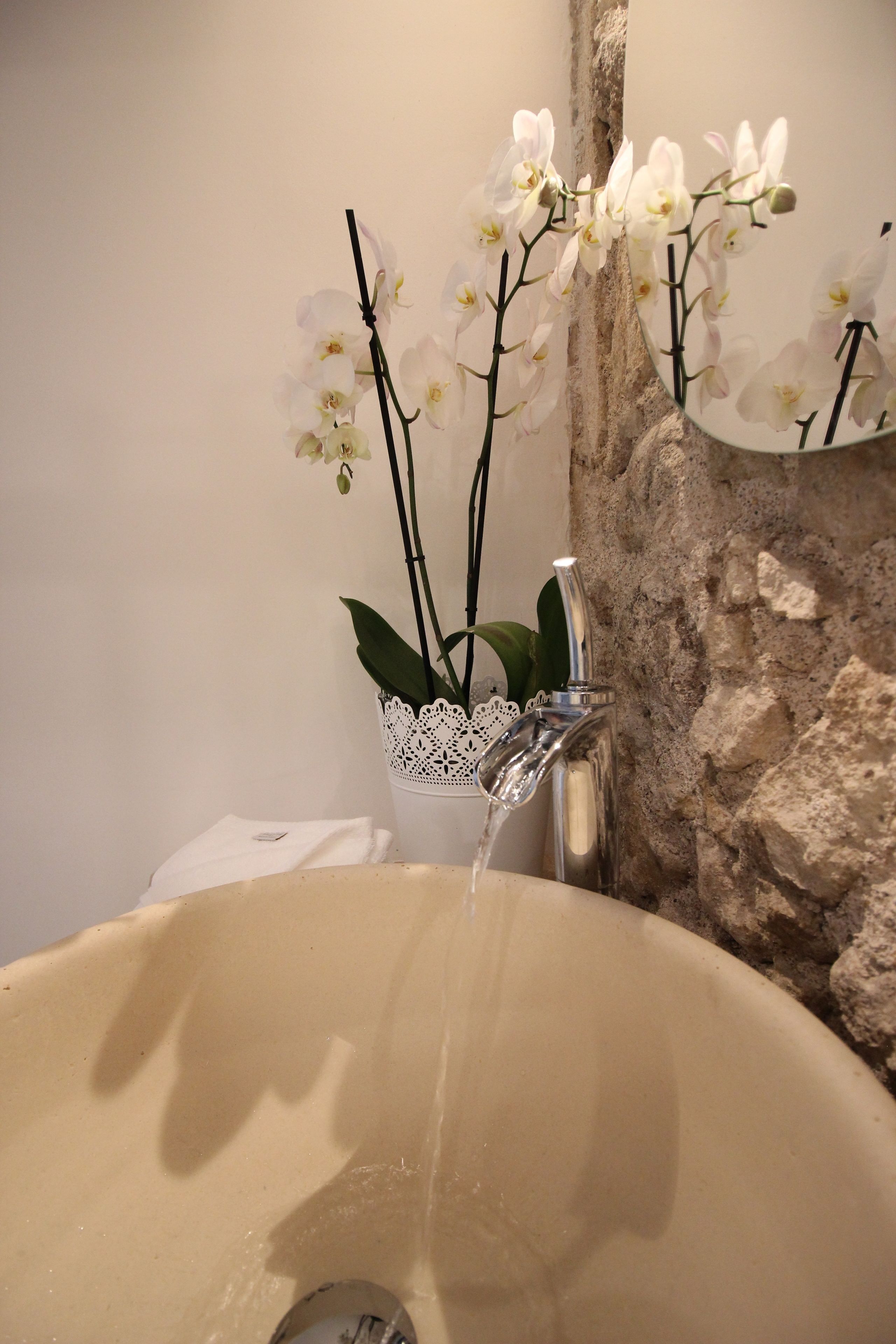 Un lavabo de salle de bain avec un mur en pierre, un récipient orné d’un motif en dentelle et des orchidées blanches.