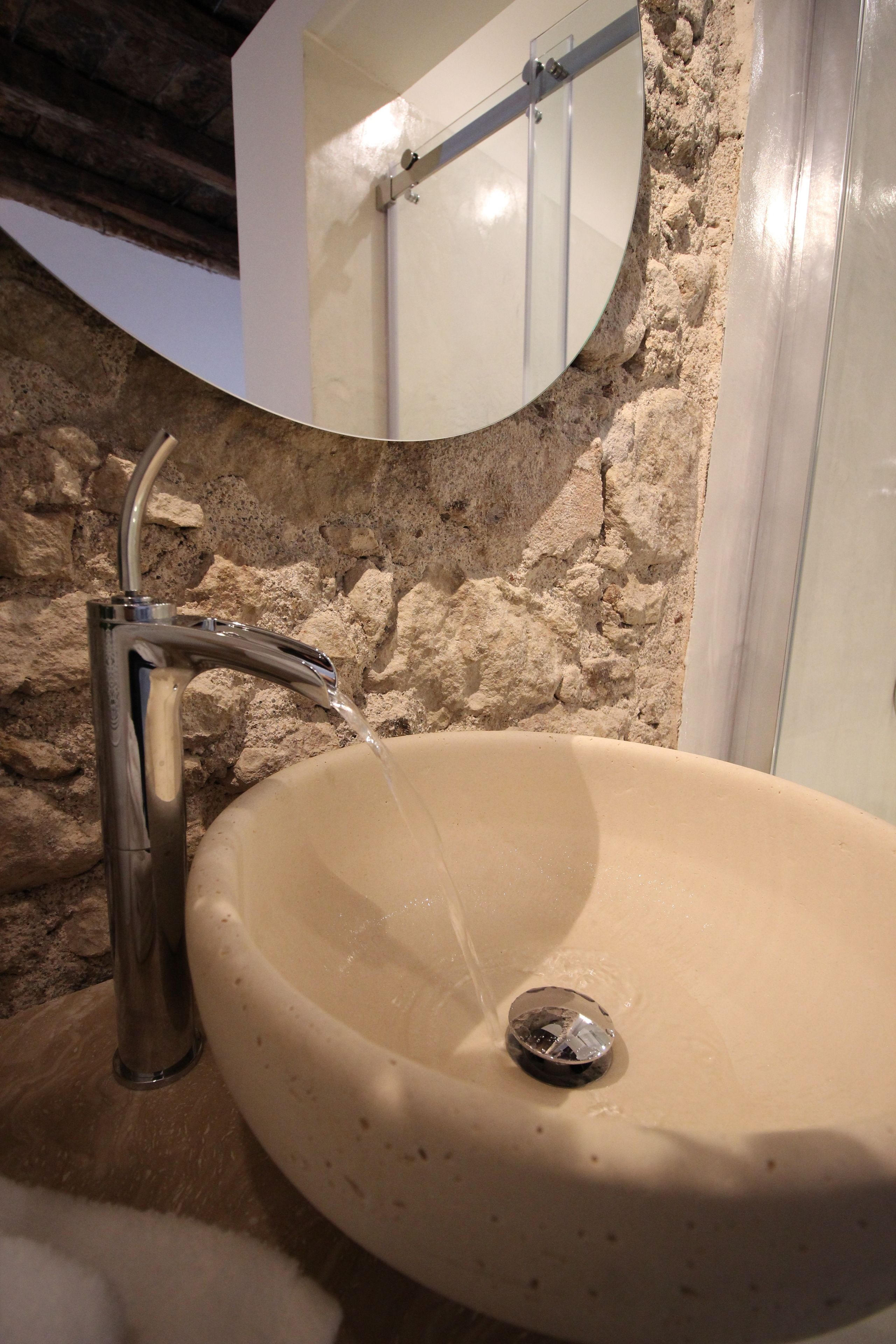 Un baño con pared de piedra, un espejo redondo y un lavabo con agua corriente.