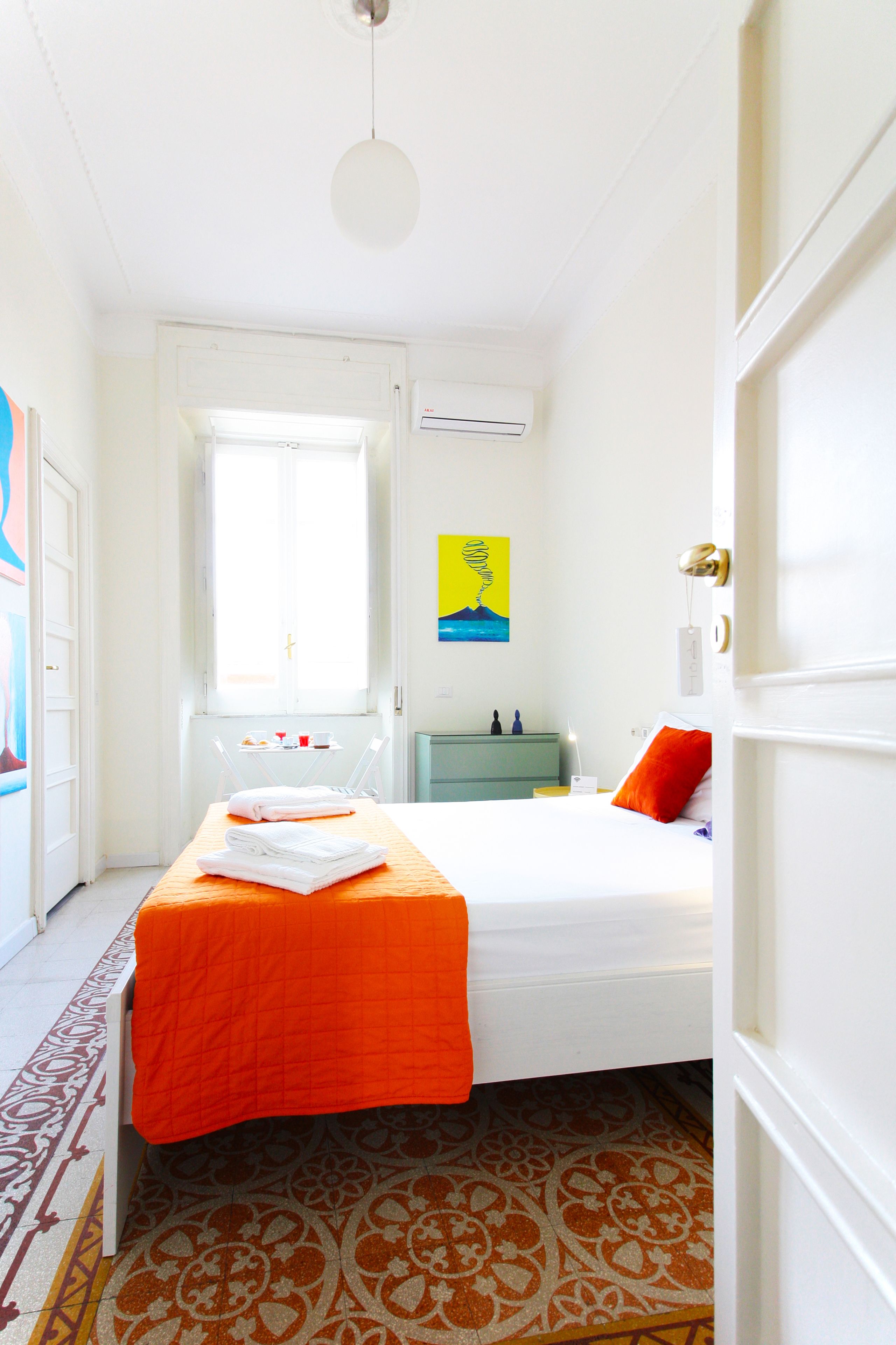 A hotel room with a bed, a window, a small table with a chair, a nightstand, and a painting on the wall.