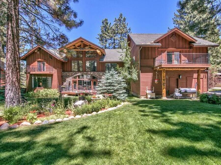 A large wooden cabin with a balcony, surrounded by a well-maintained garden and tall trees.