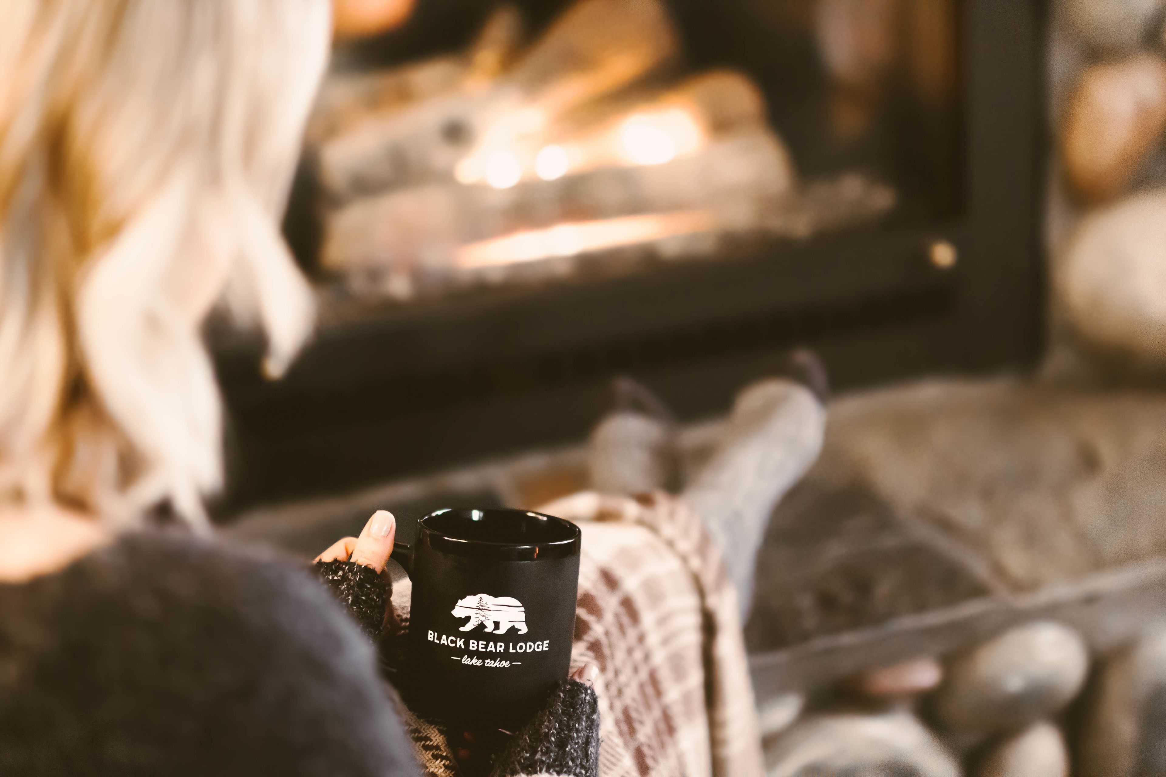 Uma pessoa segurando uma caneca com a inscrição "Black Bear Lodge", sentada em um quarto aconchegante com lareira.