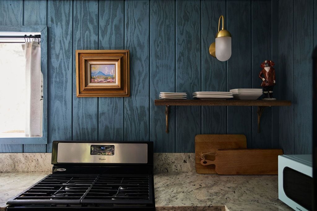 A kitchen with a stove, a microwave, a wooden shelf with dishes, a framed picture, and a hanging light fixture.