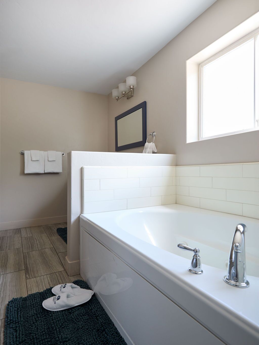 A modern bathroom with a white bathtub, a large mirror, and a window providing natural light.