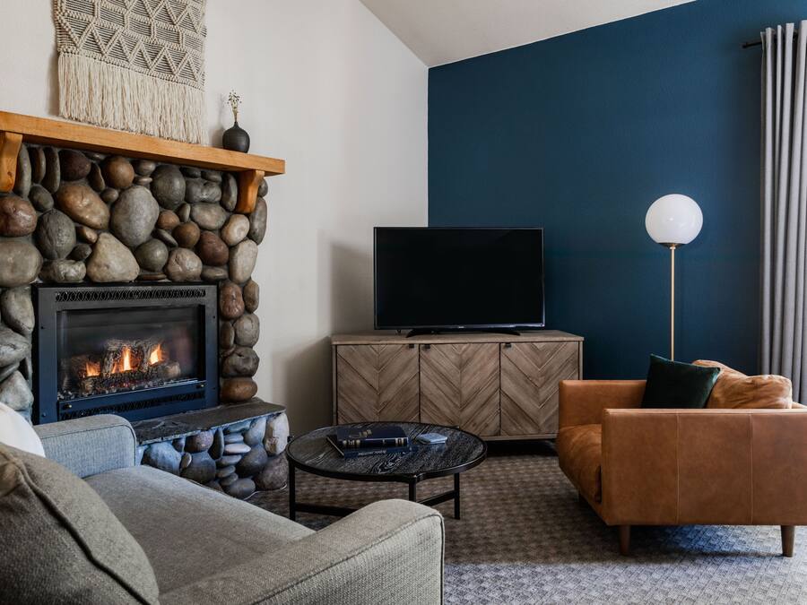 Uma sala de estar com lareira de pedra, uma televisão sobre um móvel de madeira, um sofá de couro marrom e uma mesa de centro redonda.