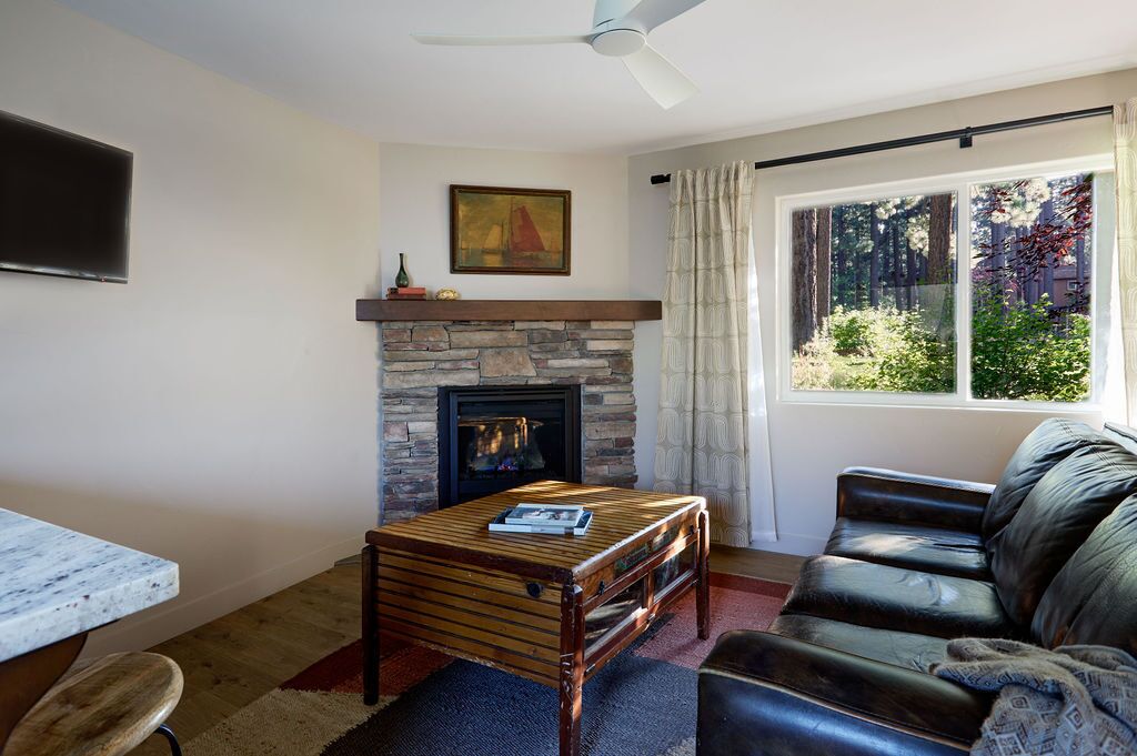 A living room with a stone fireplace, a ceiling fan, a large window with a view of trees, a leather sofa, and a coffee table.