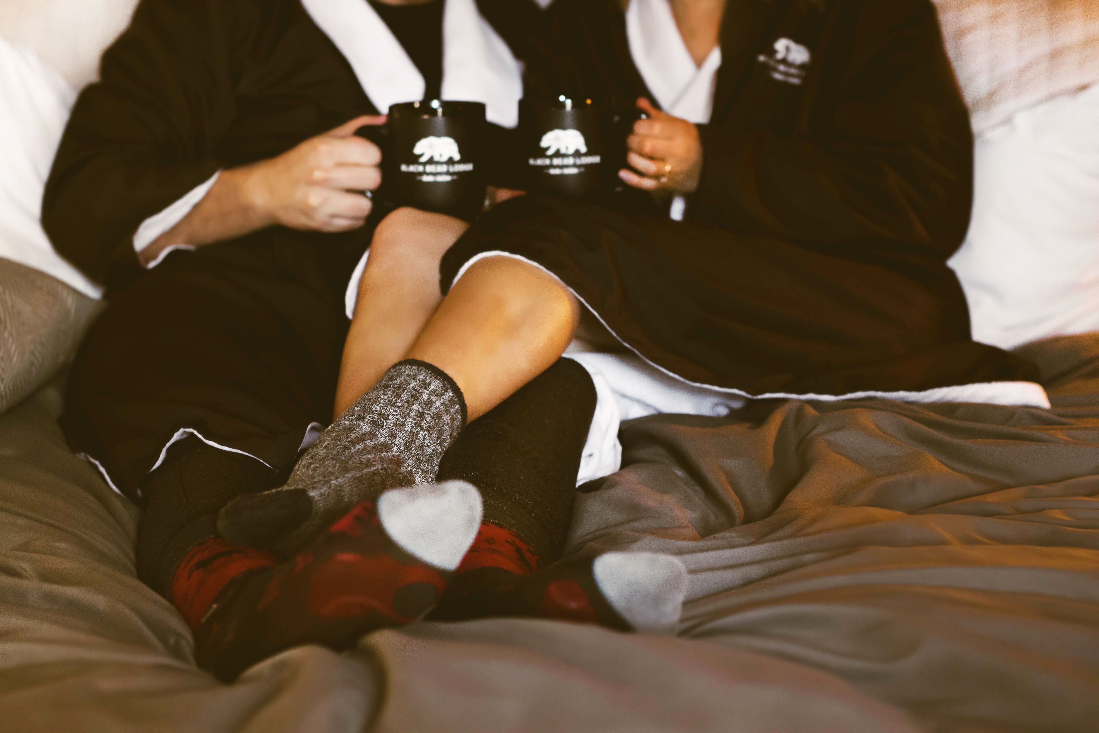Two people sitting on a bed with a blanket, holding black mugs with a white logo.