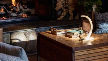A stone fireplace with a log, a fire, and a wooden wall decoration. There is a table with books and a plant.