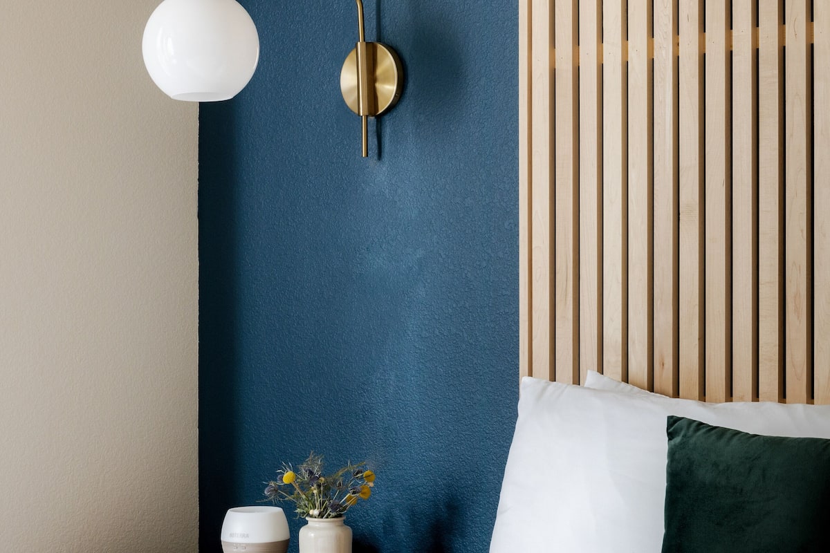 Een slaapkamer met een bed, nachtkastje met een lamp, een bloevas en een hoofdeinde van houten latten.