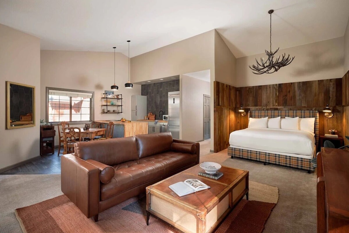 Een ruime woonkamer met een bruine leren bank, een houten salontafel en een eethoek met een houten tafel en stoelen.