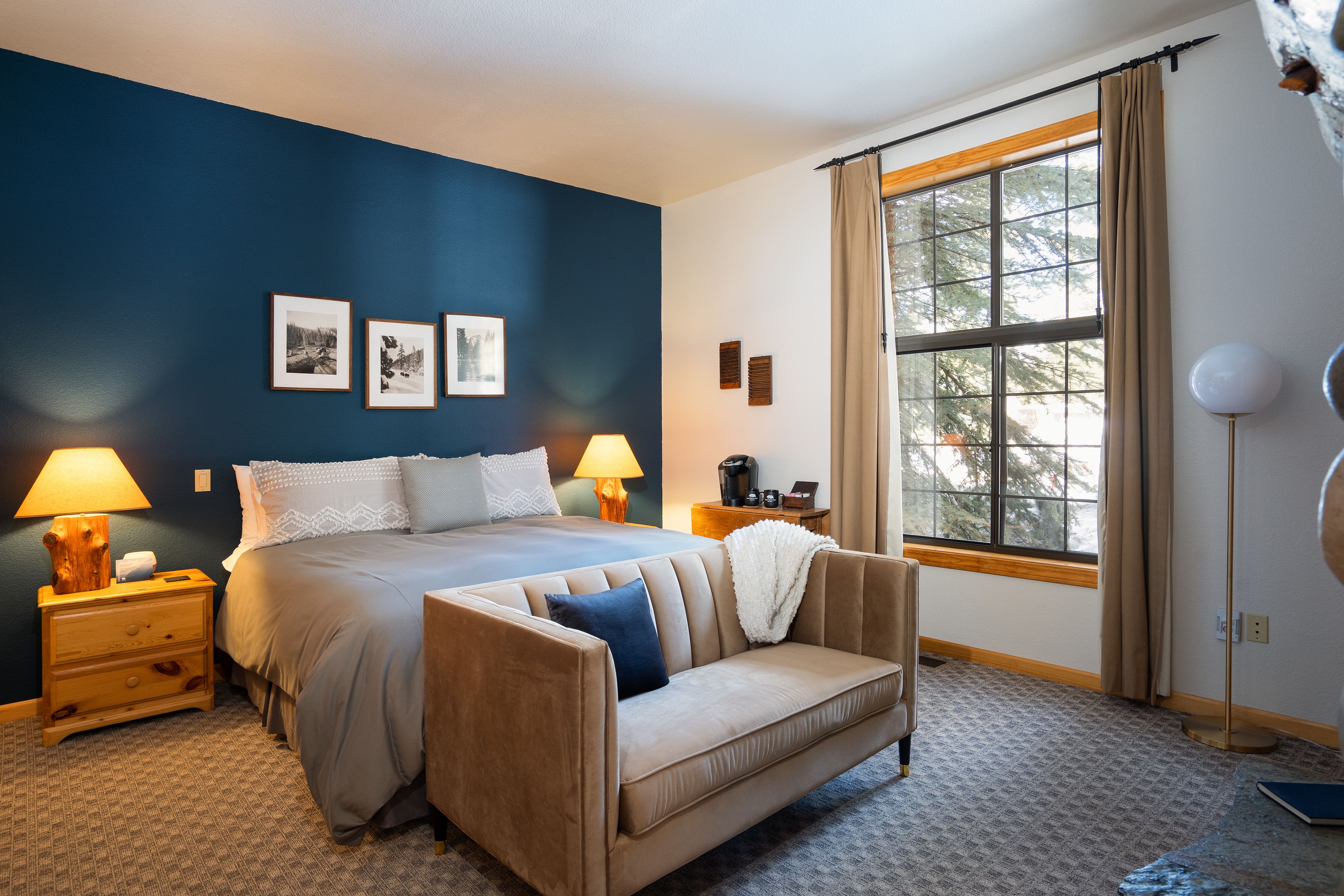 Un dormitorio con cama, sofá, mesita de noche, lámpara, ventana con cortinas e imágenes enmarcadas en la pared.