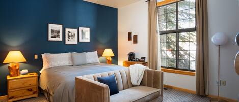 Un dormitorio con cama, sofá, mesita de noche, lámpara, ventana con cortinas e imágenes enmarcadas en la pared.