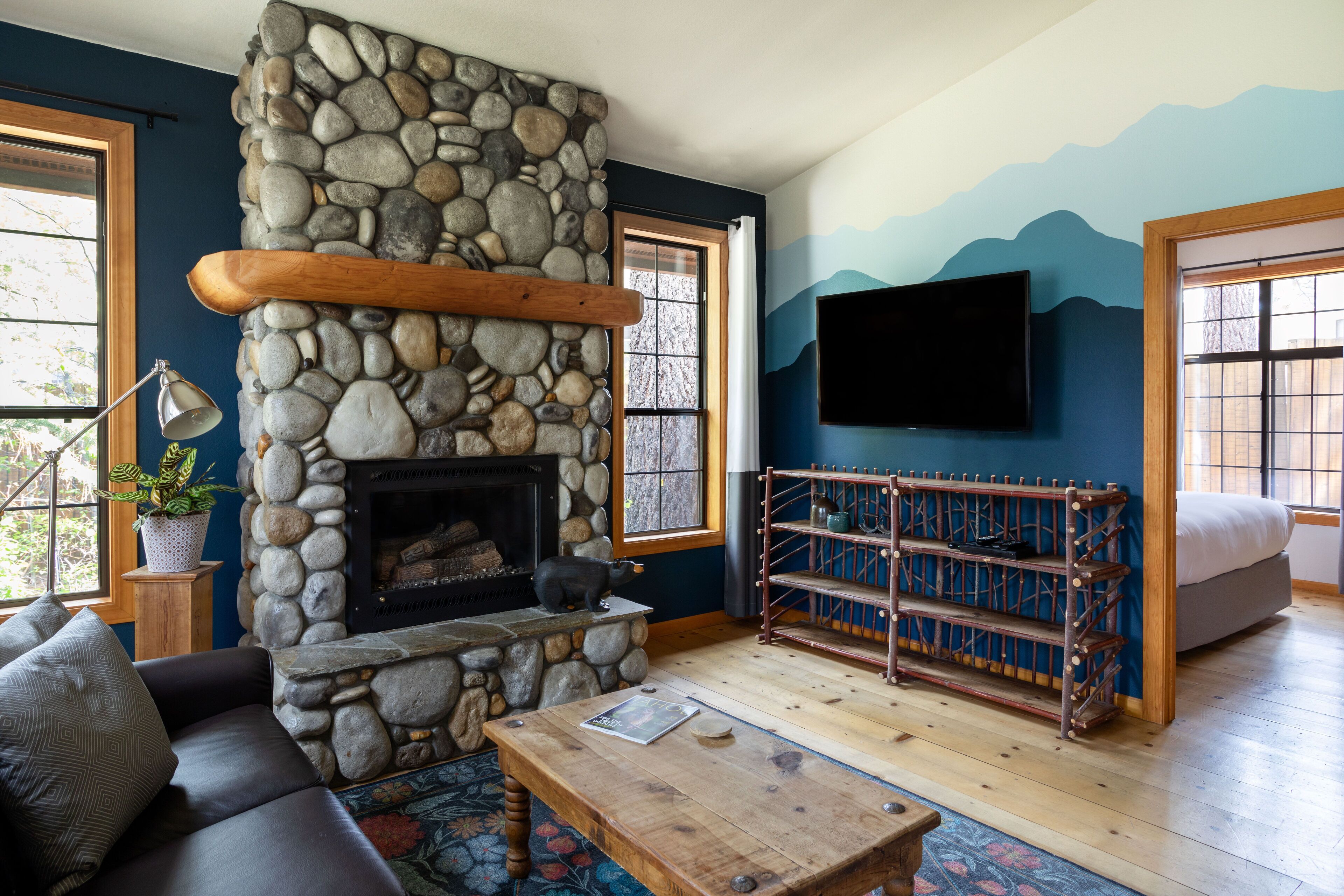 Un salon chaleureux avec une cheminée en pierre, une table basse en bois, un canapé noir et un téléviseur fixé au mur.