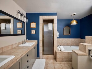 A modern bathroom with two sinks, a shower, and a bathtub. There are framed pictures on the wall, a large mirror, and a gold pendant light.