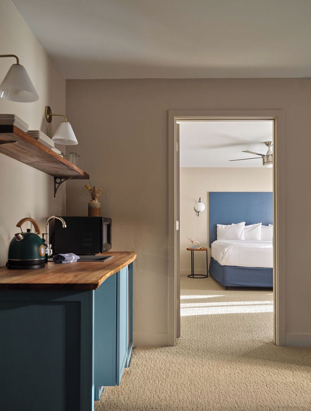 Une chambre d’hôtel avec un bureau en bois, un lit avec une tête de lit bleue, un ventilateur de plafond et une table de chevet avec une lampe.