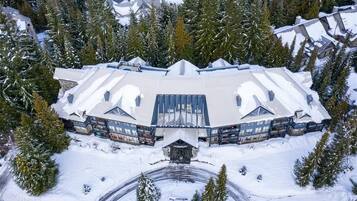 A large, snow-covered building with a symmetrical roof and multiple windows, surrounded by snow-laden trees.