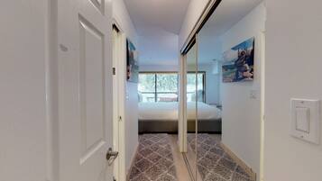 A narrow hallway leading to a bedroom with a large window, a bed, and a painting on the wall.