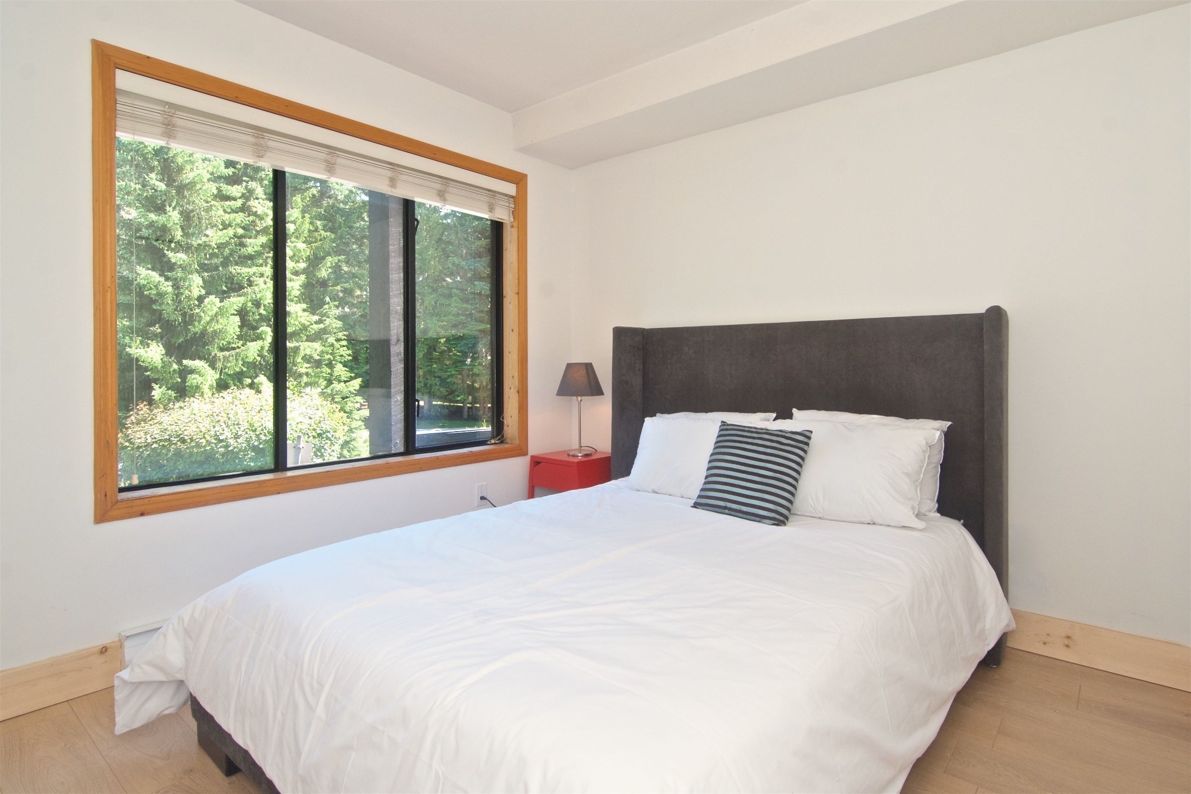 A bedroom with a large window, a bed with white bedding, a dark headboard, and a bedside table with a lamp.