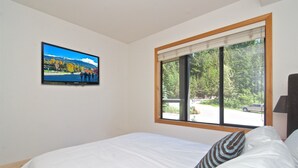 Un dormitorio con cama, televisor montado en la pared y ventana con vistas a un aparcamiento y árboles.
