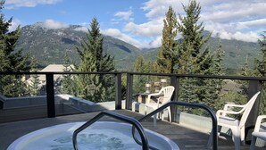 Un jacuzzi en un balcón con vistas a la montaña, rodeado de árboles y sillas de plástico blancas.