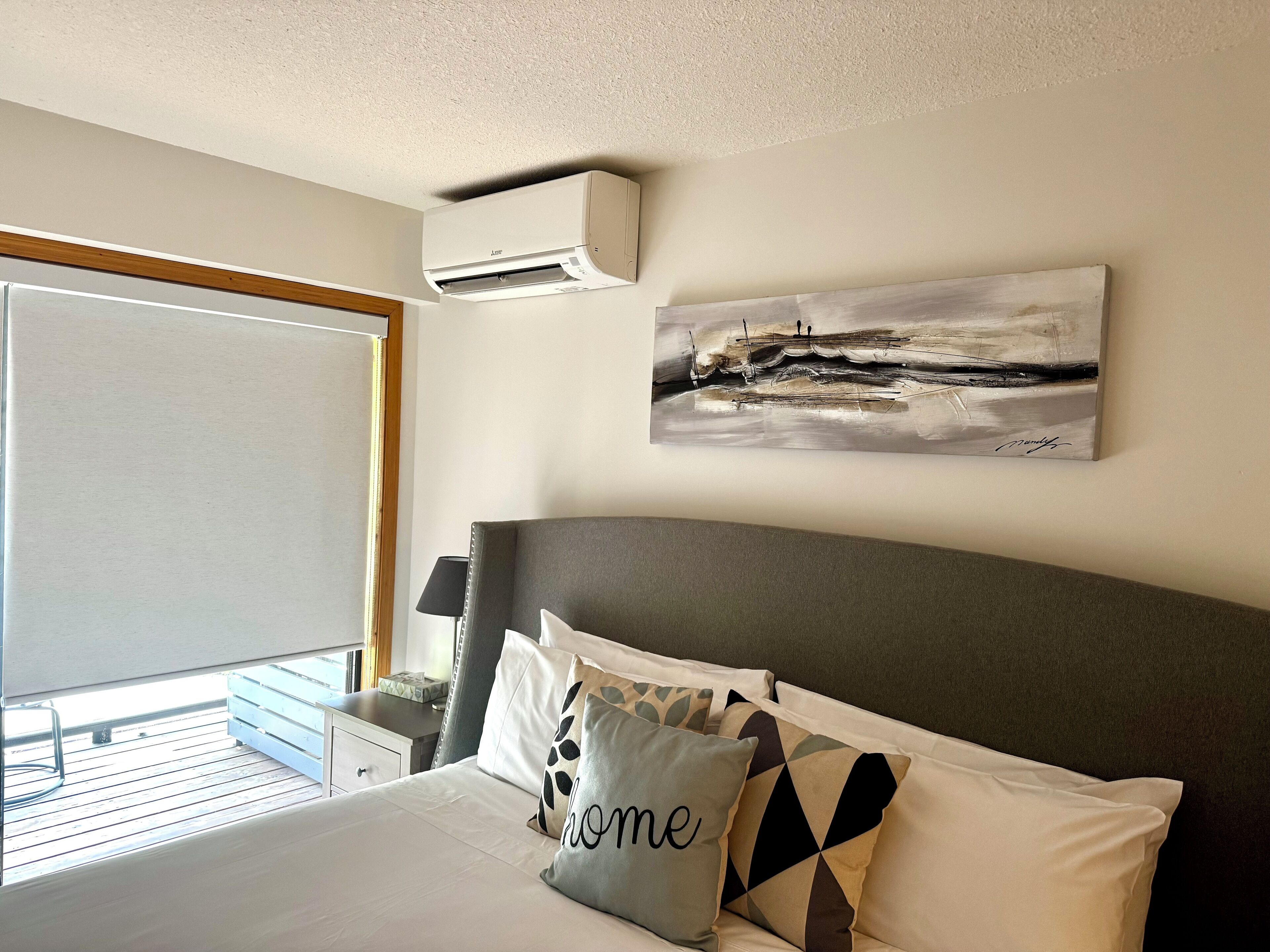 A hotel room with a bed, a painting on the wall, an air conditioning unit, and a window with a view.
