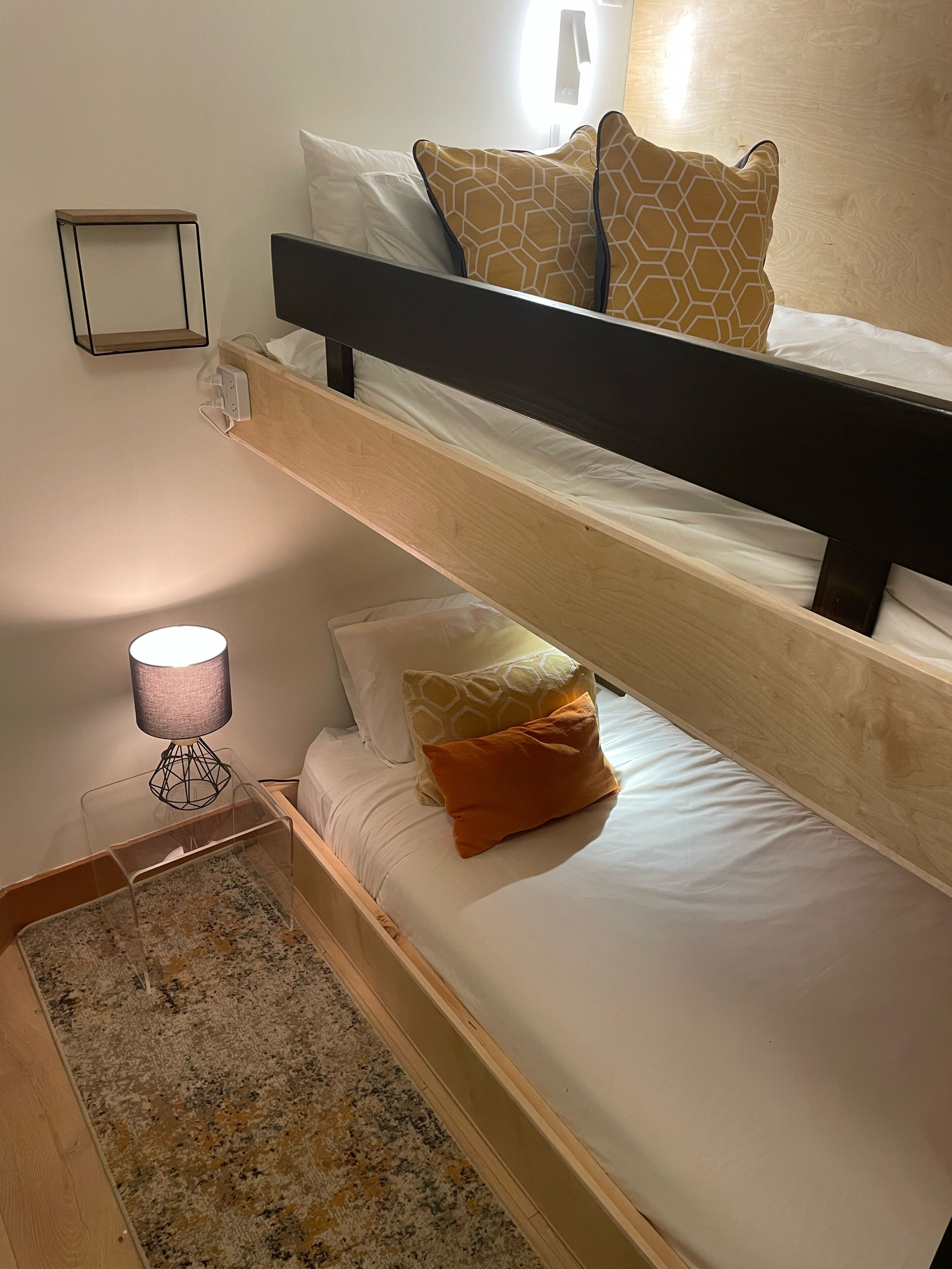 Ein Stockbett mit Holzrahmen, weißer Bettwäsche und gemusterten Kissen. Ein kleiner Tisch mit Lampe und Steckdose an der Wand.