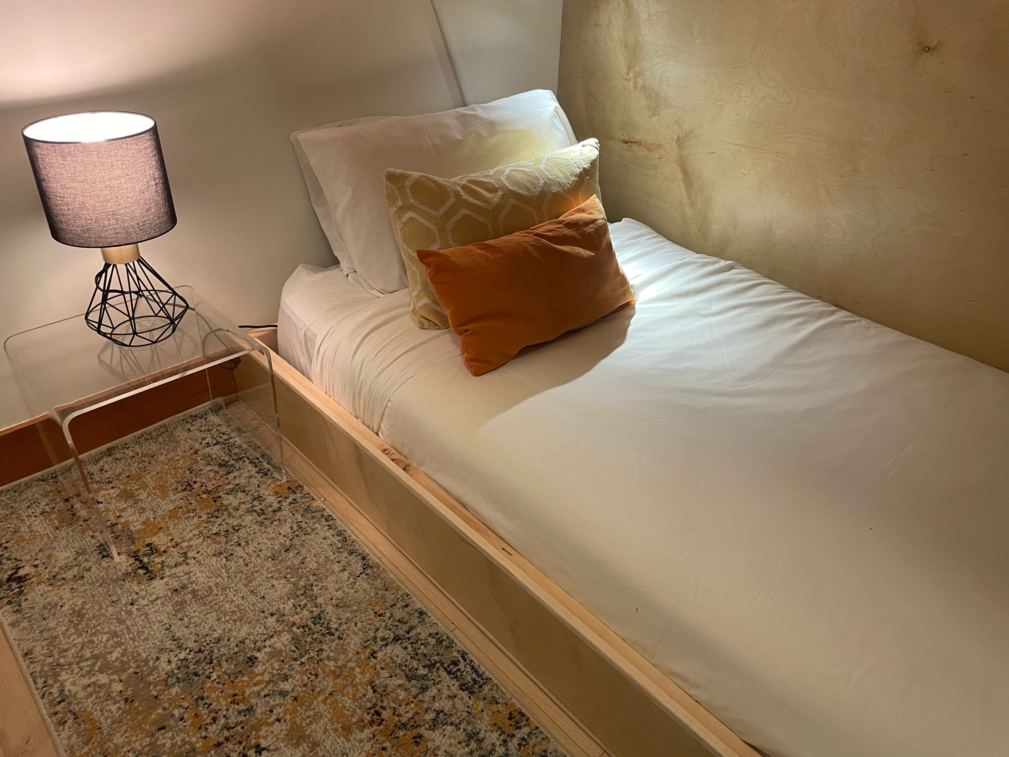 A single bed with a wooden frame, white bedding, and two pillows, one patterned and one solid orange. A lamp with a geometric base is on a clear stand beside the bed.