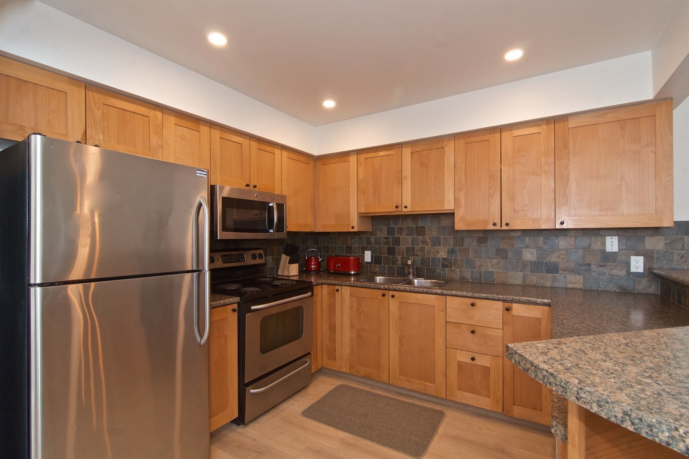 A modern kitchen with wooden cabinets, stainless steel appliances, a granite countertop, and a stone backsplash.