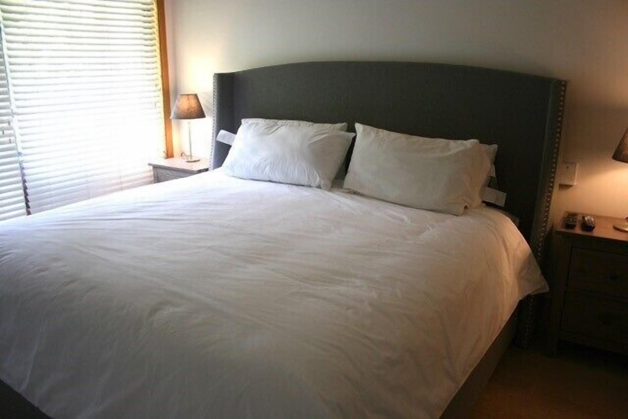 Una cama bien tendida con sábanas y almohadas blancas, una mesita de noche con una lámpara y una ventana con persianas.