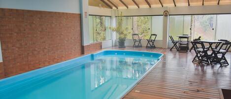 A swimming pool with wooden decking, a covered patio with chairs, and large windows.