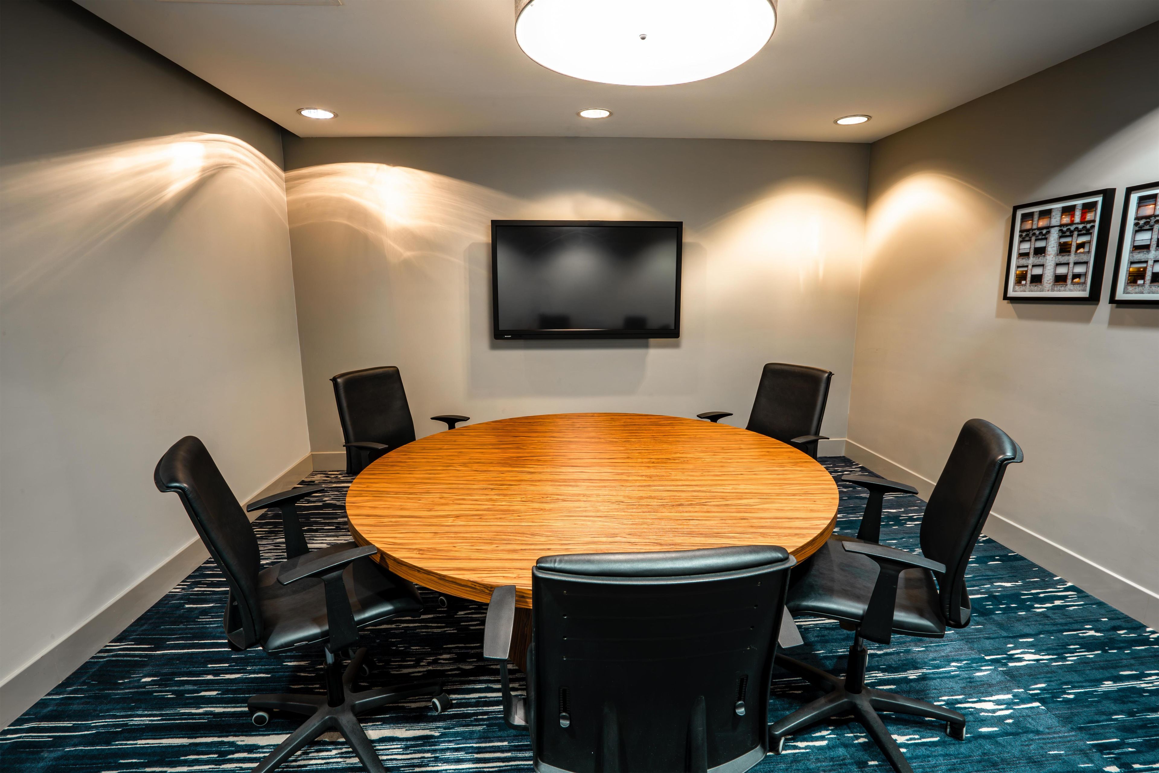A conference room with a round wooden table, six black chairs, a flat-screen TV on the wall, and patterned carpet.