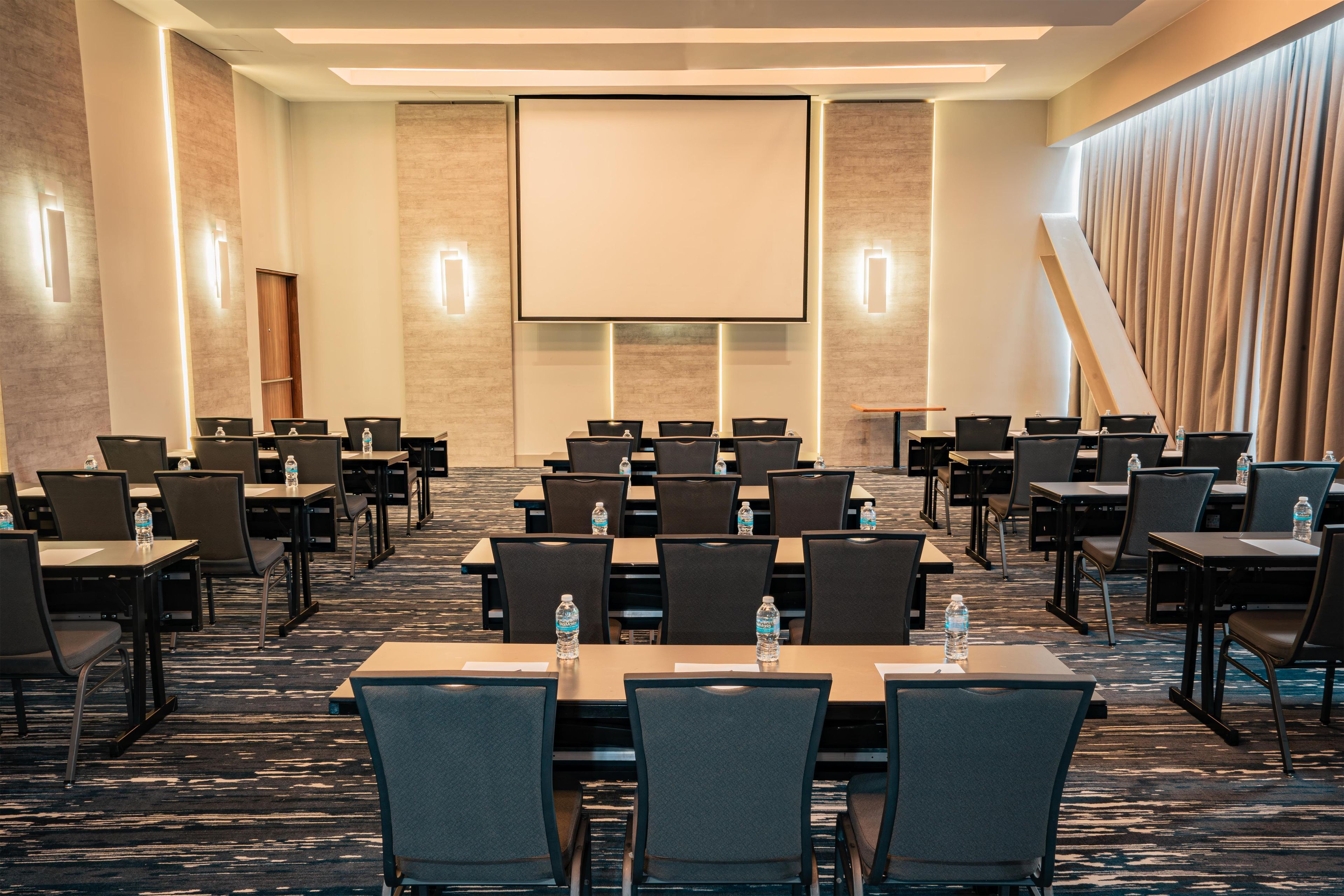 A conference room with rows of tables and chairs, a large screen at the front, and a water bottle on each table.