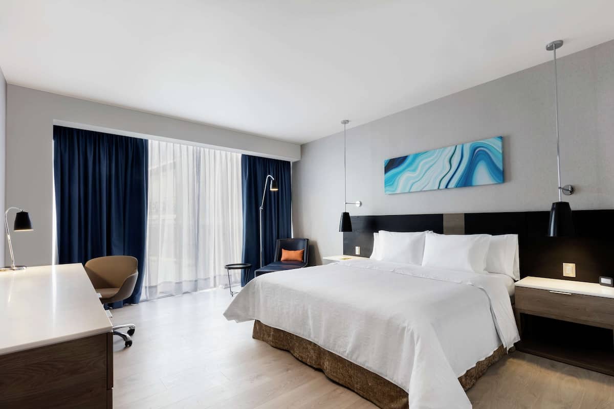 A modern hotel room with a large bed, a desk, a chair, and a view of the outside through large windows.