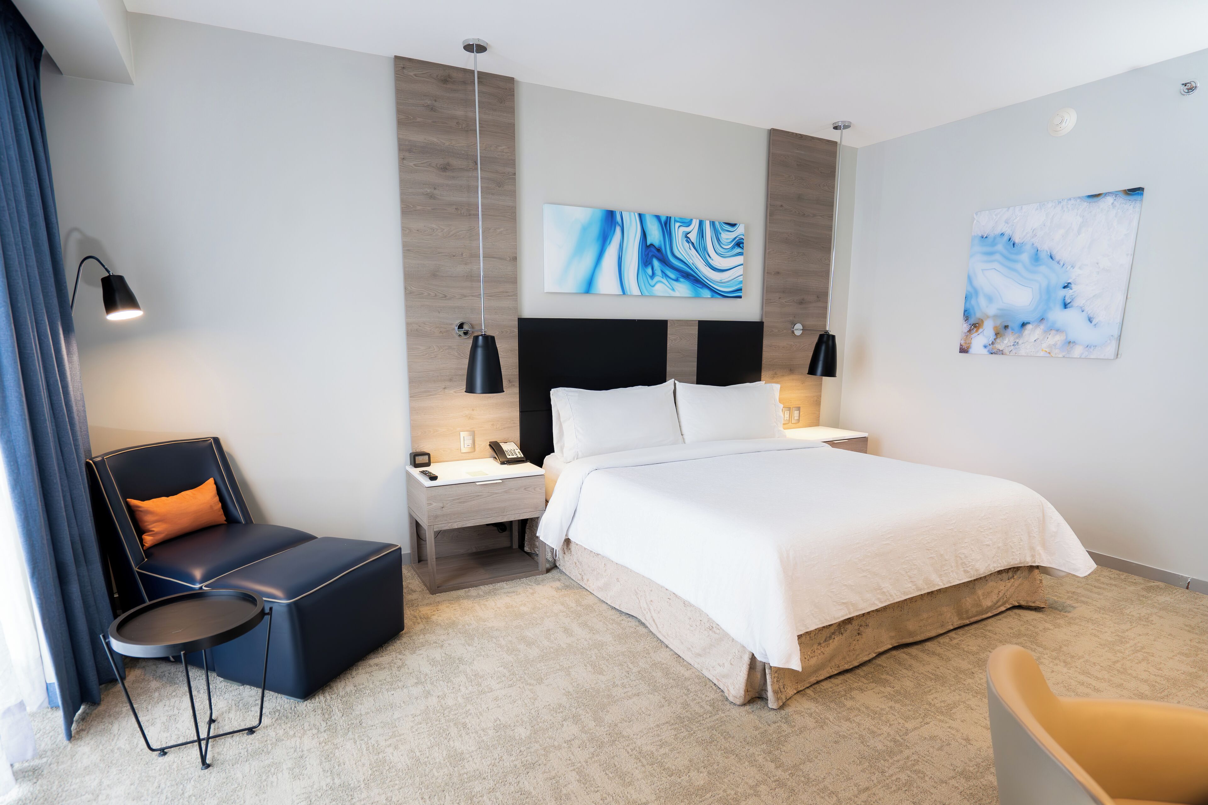 Quarto de hotel moderno com uma cama grande, uma mesa de cabeceira, uma cadeira e obras de arte nas paredes.