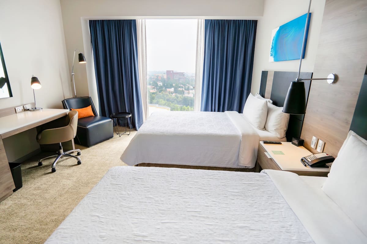 A hotel room with two beds, a desk, a chair, a lamp, and a view of the city.