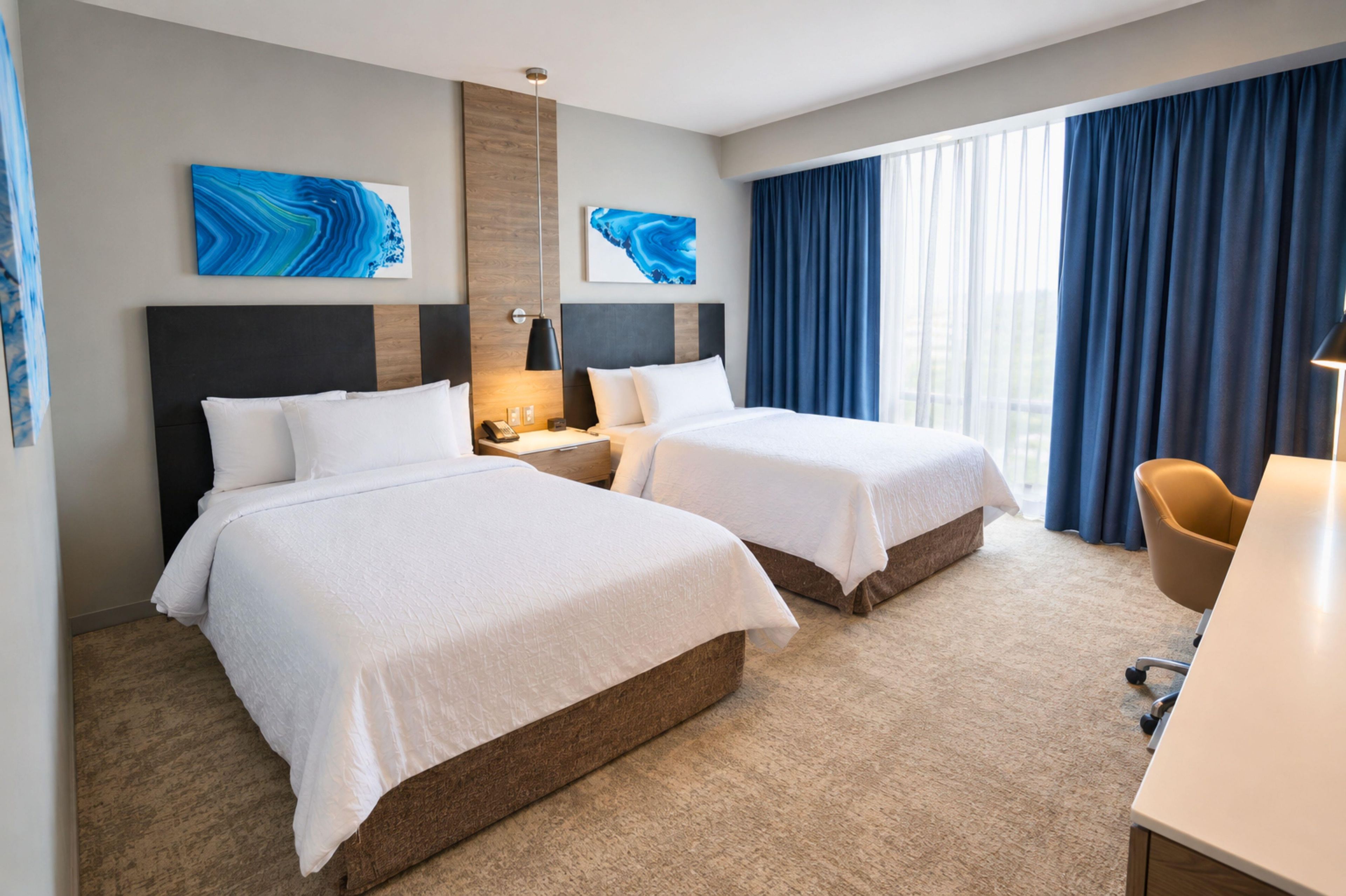 A hotel room with two beds, a desk, a chair, and artwork on the walls.