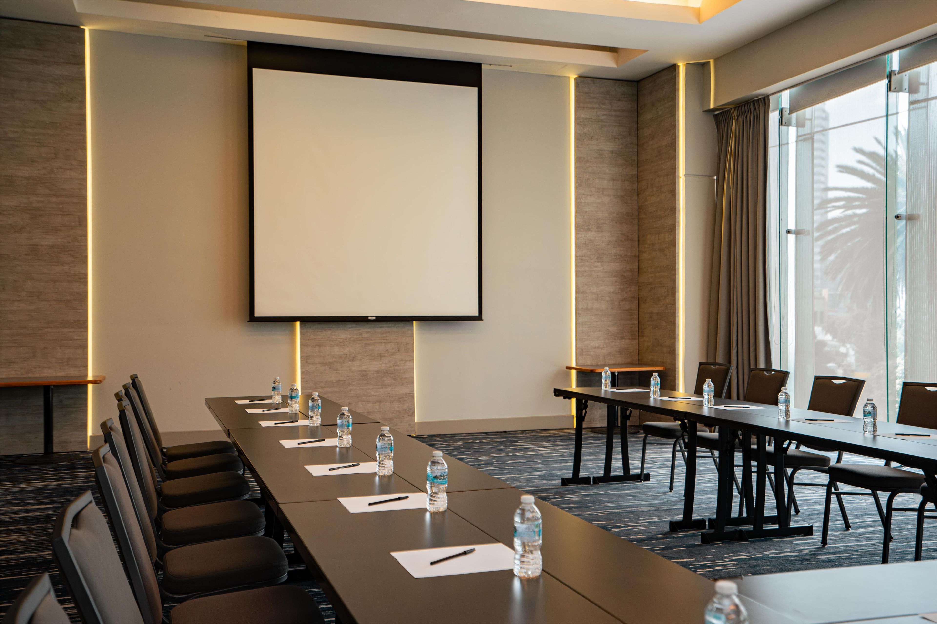 A conference room with a long table, chairs, water bottles, and a projector screen.
