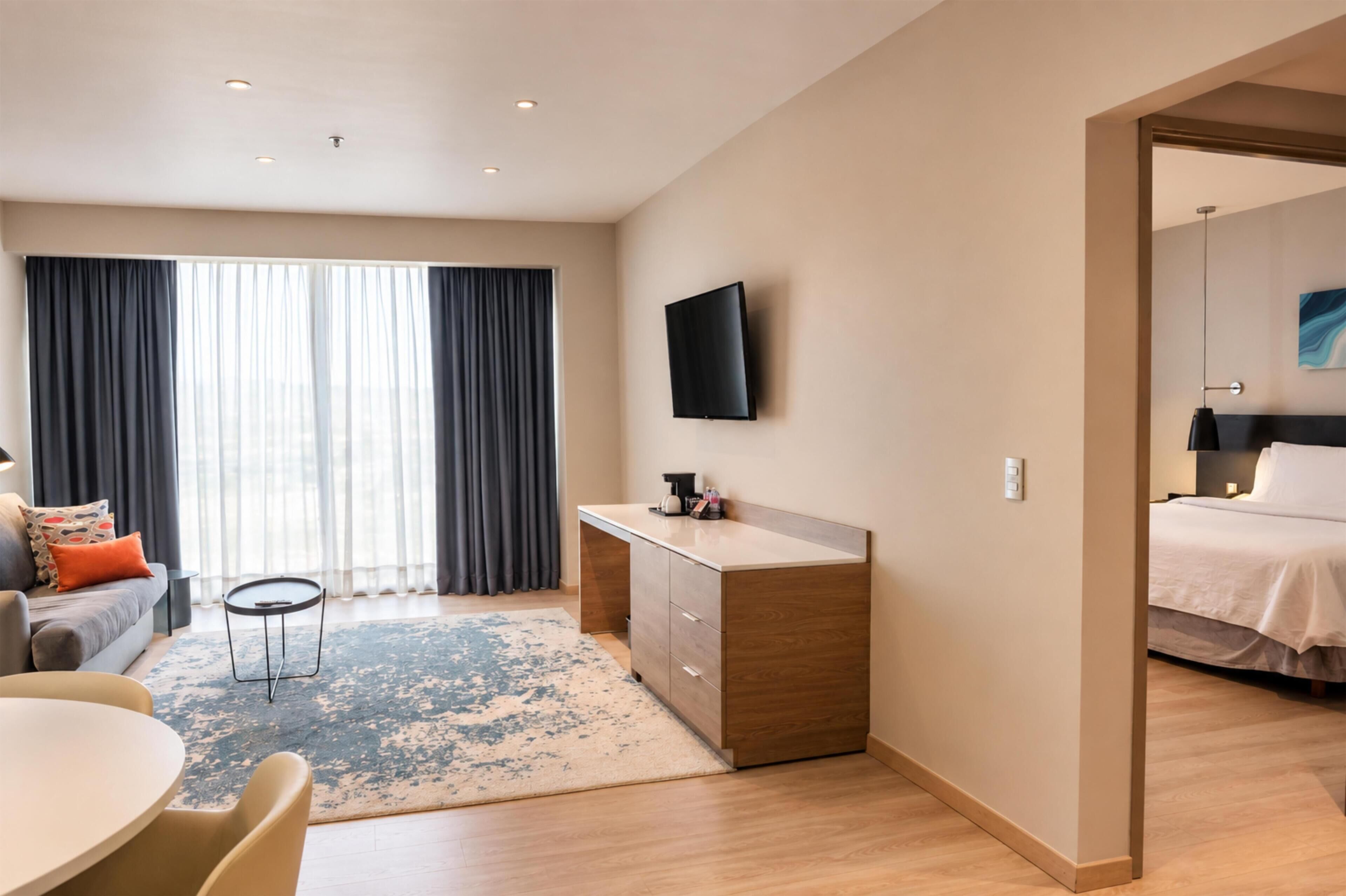 A modern hotel room with a flat-screen TV, a sofa, a dining table, and a separate bedroom area.