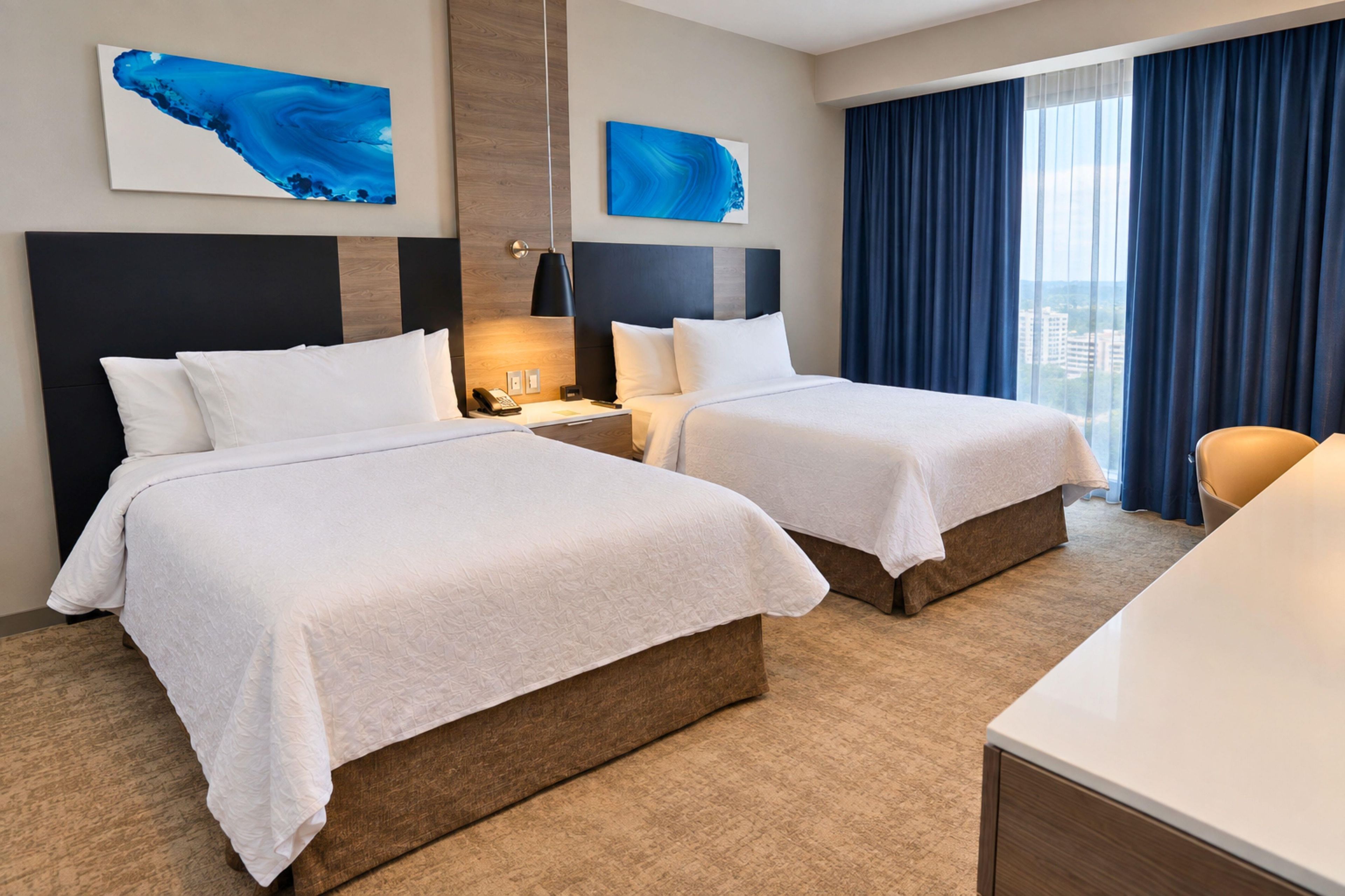 A hotel room with two beds, a desk, and a view of the city.