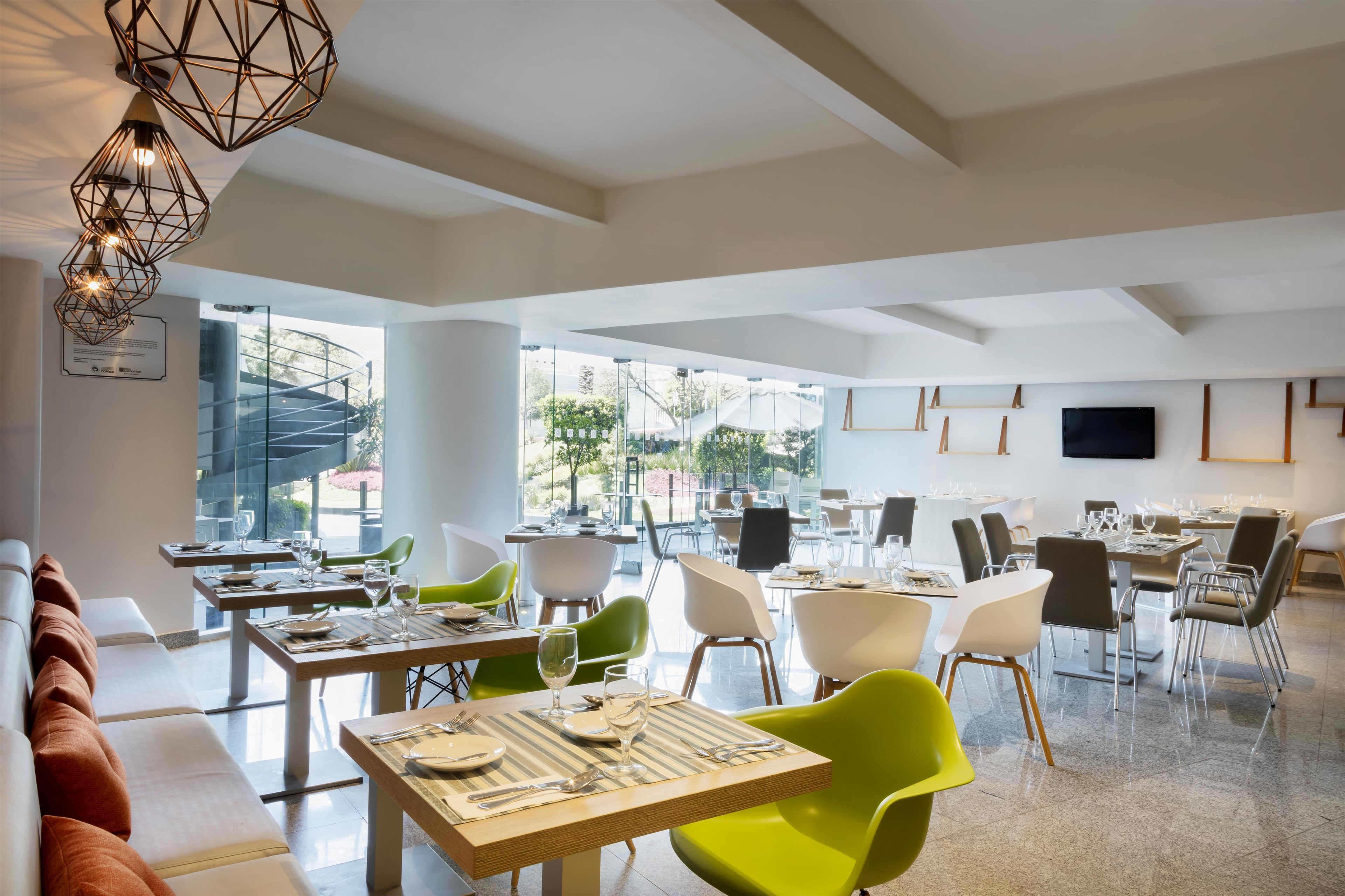 A modern dining area with multiple tables and chairs, large windows offering a view of the outdoors, and a geometric ceiling light fixture.