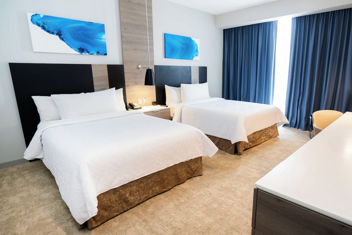 A hotel room with two beds, a desk, and a chair. There are two paintings on the wall and a window with curtains.