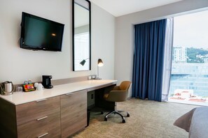 Una habitación de hotel con escritorio, silla, televisor y un ventanal con cortinas.