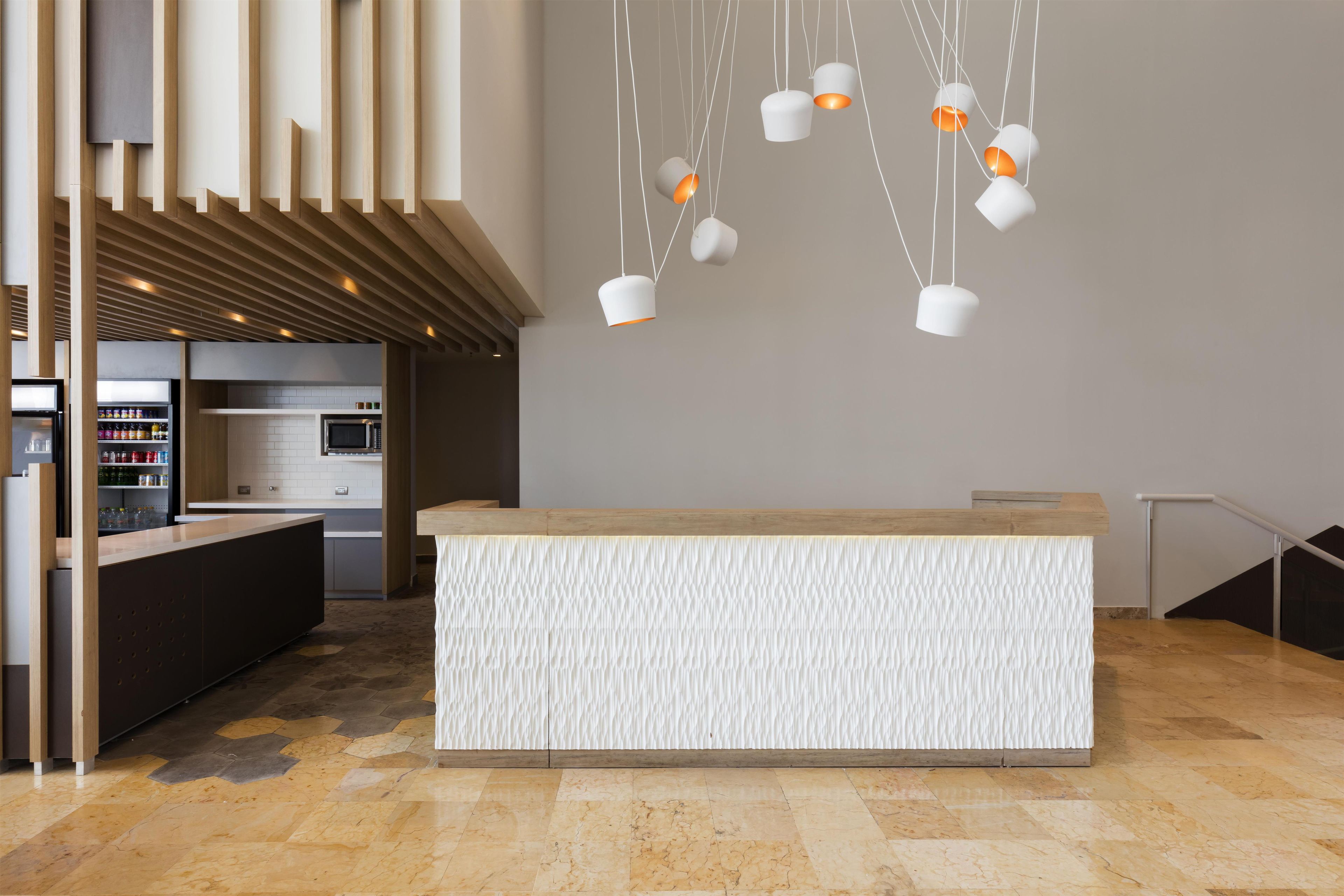 A modern reception desk with a textured white surface, a kitchen area with a microwave and refrigerator, and pendant lighting with white and orange shades.